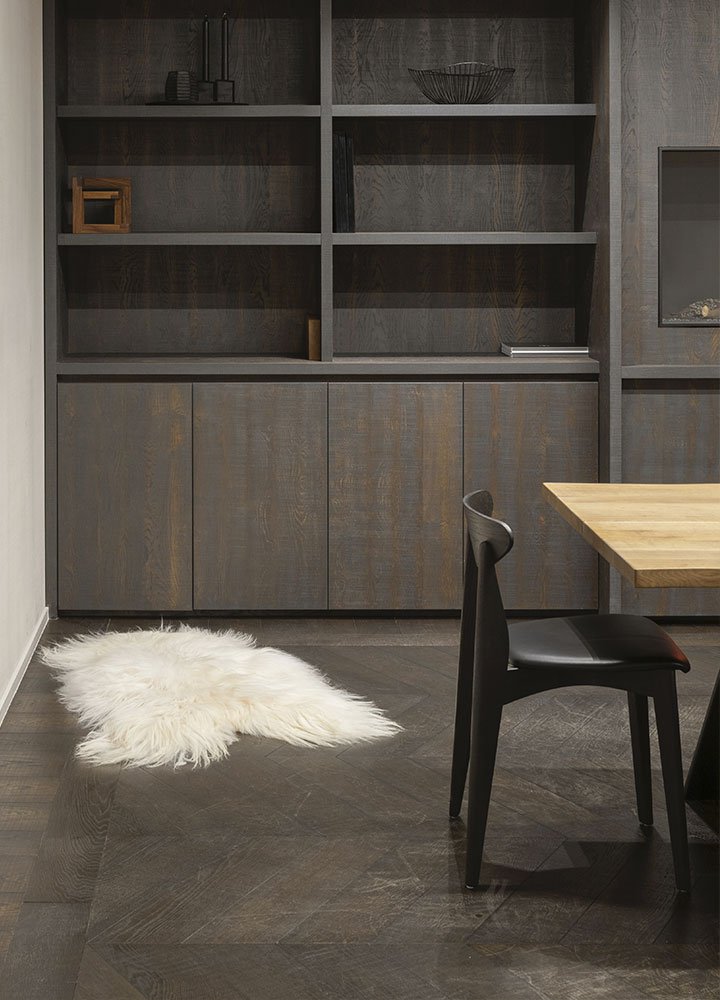 Dining room dominated by dark wood tones with a chair, part of a rectangular table, a bookshelf in the background and a white pile carpet.